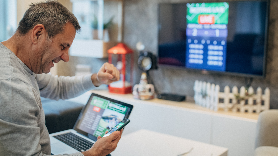 Man seated at home, navigating furlough claims on his phone and laptop, with a TV screen displaying a sports betting app interface in the background.