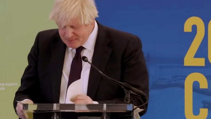 Boris Johnson, with his distinctive blonde hair, stands behind a clear podium reading papers. Dressed in a suit and tie, he speaks into the microphone positioned before him. A blue and yellow backdrop frames the scene.