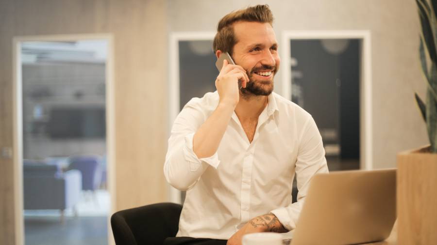 A man in a white shirt is smiling while talking on his phone and working on a laptop, seated in a modern office environment, likely discussing HR strategies or personnel management.