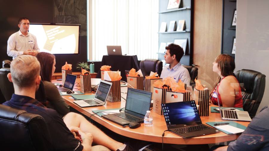 A person is giving a presentation to a group of six people seated around a conference table, discussing key People Management strategies. Laptops, notebooks, and gift bags are on the table. A large screen displaying an HR slide is in the background.