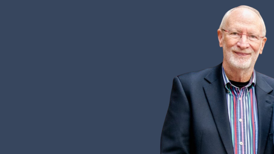 Elderly man with glasses and a white beard wearing a dark blazer over a colorful striped shirt, standing against a plain dark background, smiling lightly despite his past struggle with work addiction.