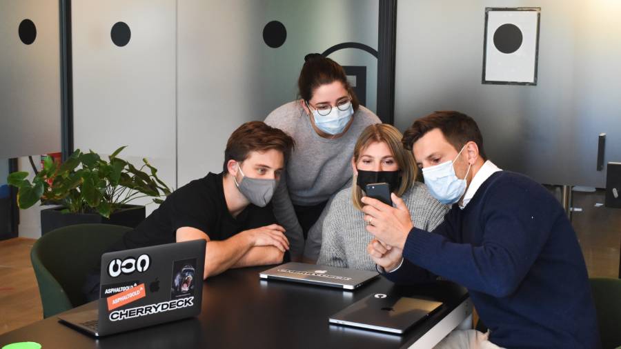 A group of four people wearing masks, part of the vaccination workforce, gathered around a table with laptops, looking closely at a smartphone.