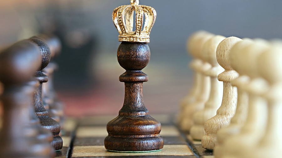 Close-up of a chessboard with a dark pawn in the center wearing a gold crown, symbolizing leadership in people management, surrounded by both dark and light chess pieces in two rows.
