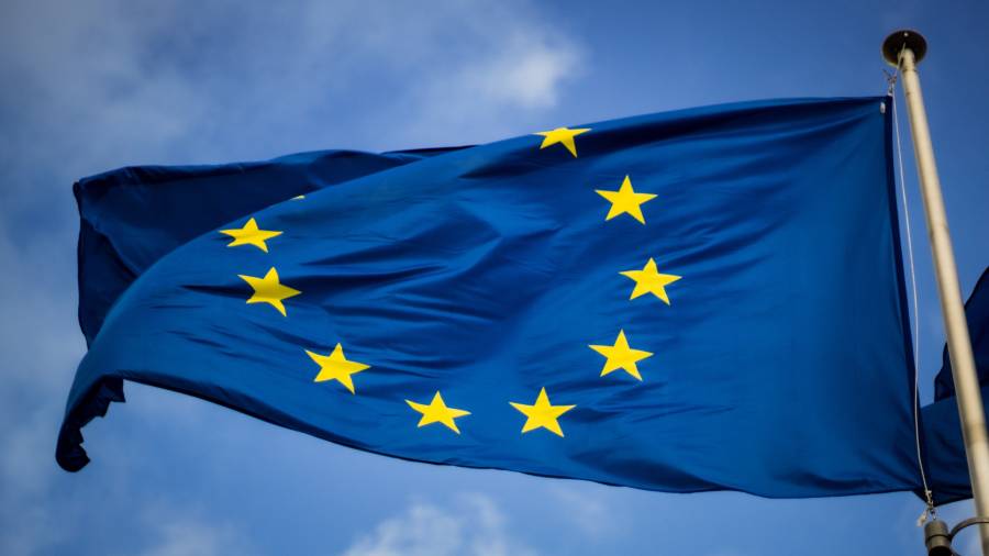 Blue flag with twelve yellow stars arranged in a circle, representing the flag of the European Union, waving against a backdrop of a clear blue sky with some clouds—symbolizing unity and people management across diverse nations.