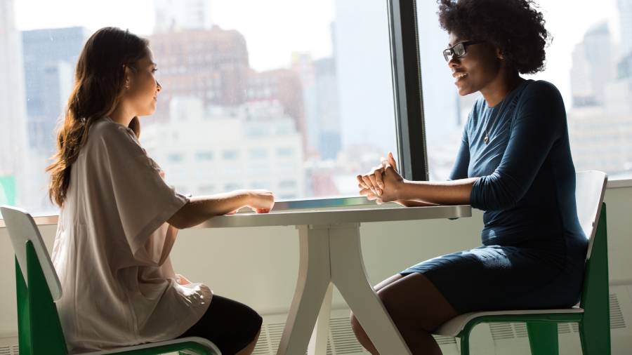 Two women are sitting across a small table in a brightly lit room, engaged in a conversation about Human Resources. The background features large windows showing an urban cityscape.