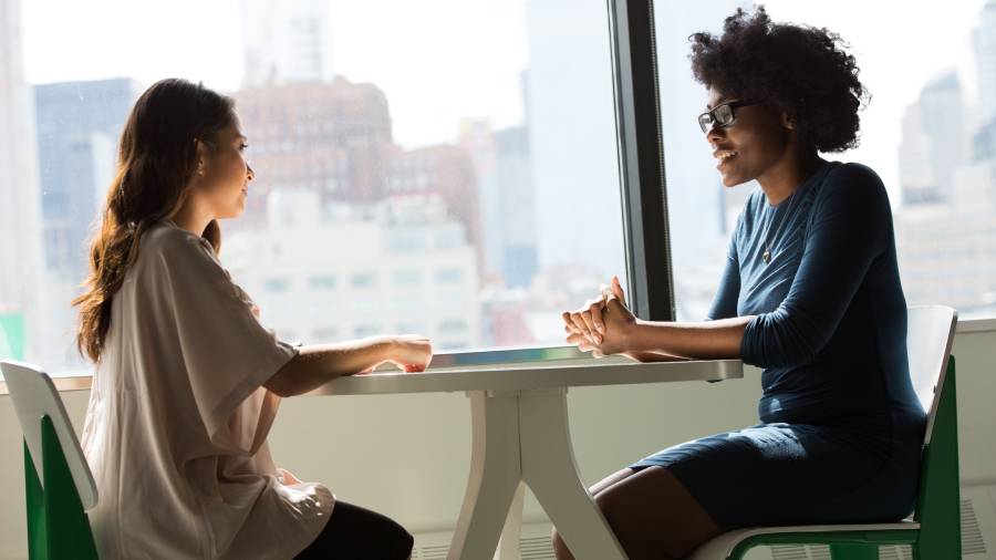 Two individuals sit across from each other at a table in front of a large window, engaged in an HR discussion. The urban skyline is visible in the background.
