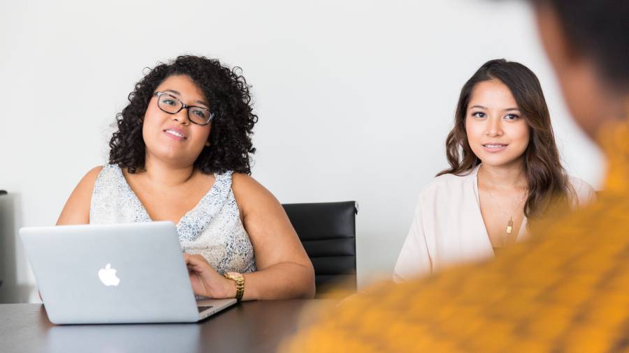 Two people are sitting at a table engaging in a conversation, likely about people management. One is using a laptop, while the other listens attentively. A third person with blurred details is partially visible in the foreground, possibly part of the HR personnel. Two people are sitting at a table engaging in a conversation, likely about people management. One is using a laptop, while the other listens attentively. A third person with blurred details is partially visible in the foreground, possibly part of the HR personnel.