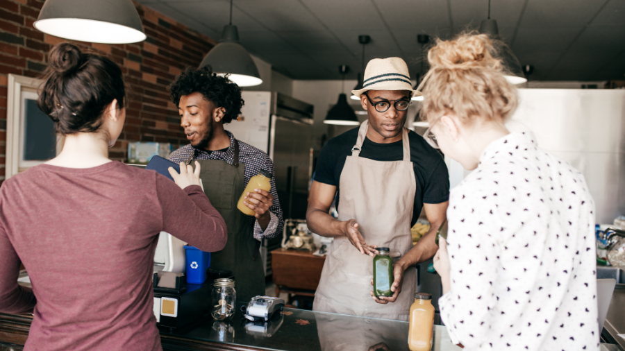 Two men wearing aprons serve two women at a juice bar in a cozy, casual atmosphere. One man confidently holds an orange juice, and the other offers a green juice while the women place their orders, reminiscent of the organized efficiency often seen in government operations.