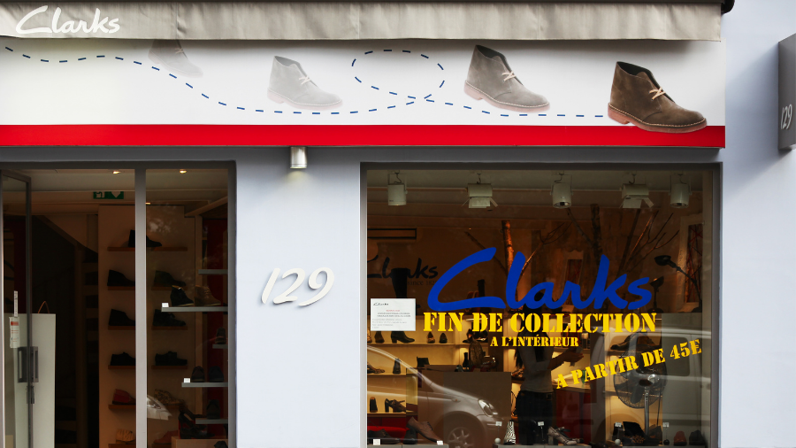Storefront of a Clarks shoe store with a display advertising the Clarks Strike end-of-collection sale starting at 45€. Shoes are visible in the window and on the signage above the entrance.