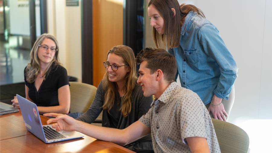 Four people are gathered around a table, looking at a laptop screen. One person is pointing at the screen, while the others watch attentively, demonstrating strong employee loyalty as they collaborate closely on the project.