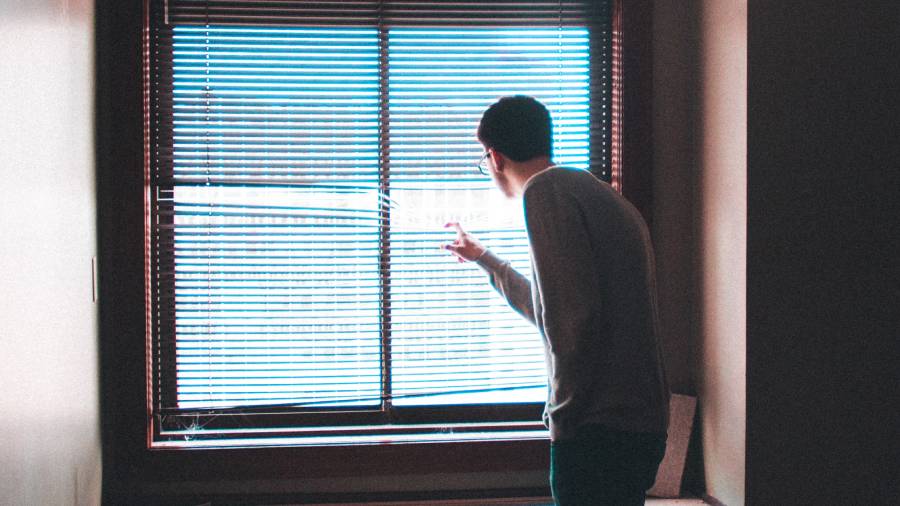 A person wearing glasses stands near a window with partially drawn blinds, looking outside and pointing with one hand, as if indicating something of interest to the HR department.