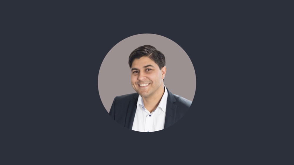 A man with dark hair is smiling, wearing a dark suit jacket over a white shirt. The background is dark grey, subtly underscoring the confidence of someone embracing a four-day week.