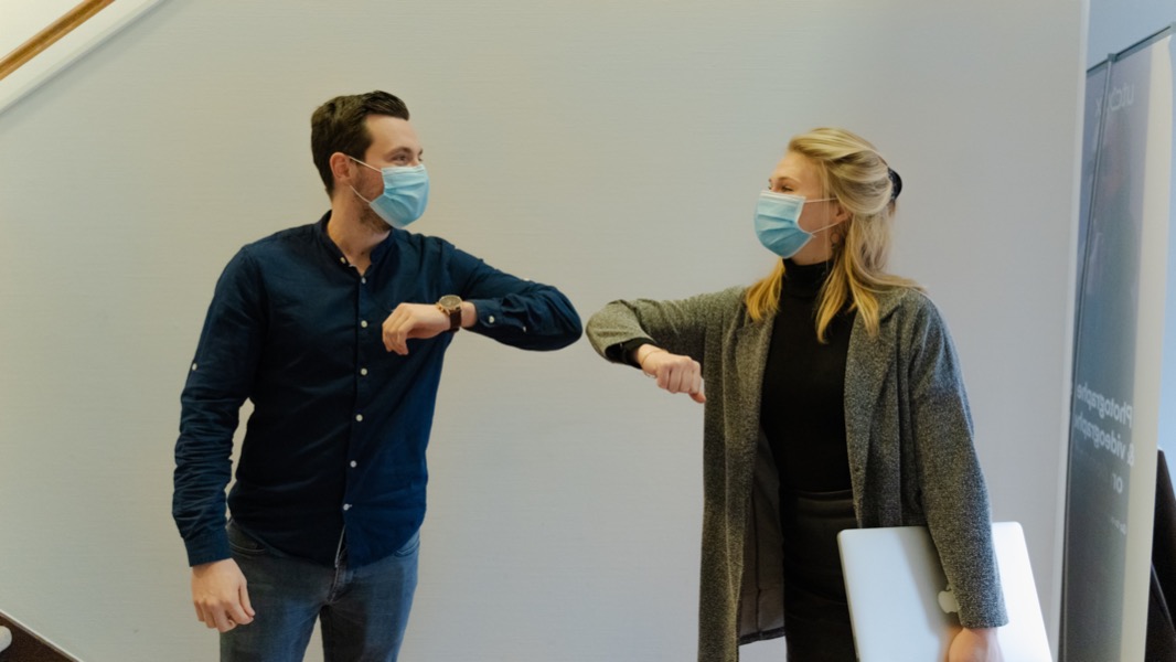 Two people wearing masks greet each other by touching their elbows inside a building, reflecting the cautious optimism of the job market. One holds a laptop, and both appear to be smiling behind their masks.