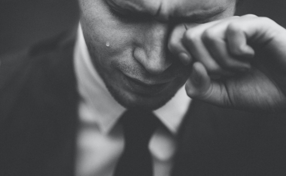 Close-up black and white image of a person in a suit wiping a tear from their eye, appearing distressed, highlighting the importance of stress management in the workplace.