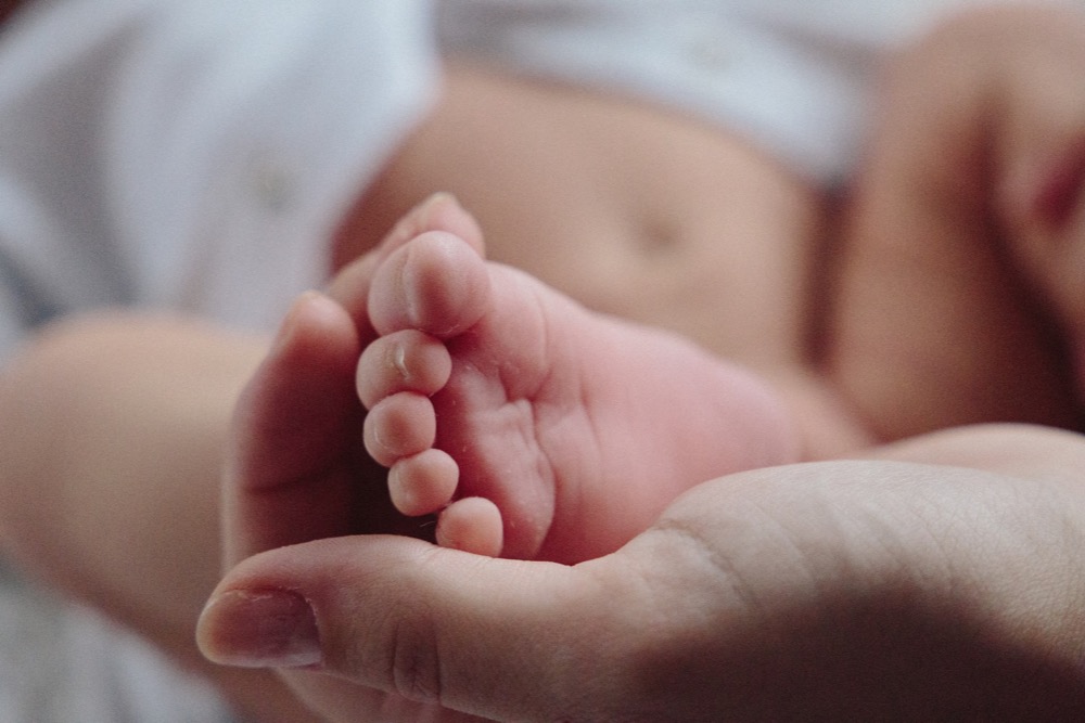A close-up of an adult hand gently cradling an infant's foot, embodying the balance and tenderness akin to flexible working. The infant is lying down, partially clothed in white.