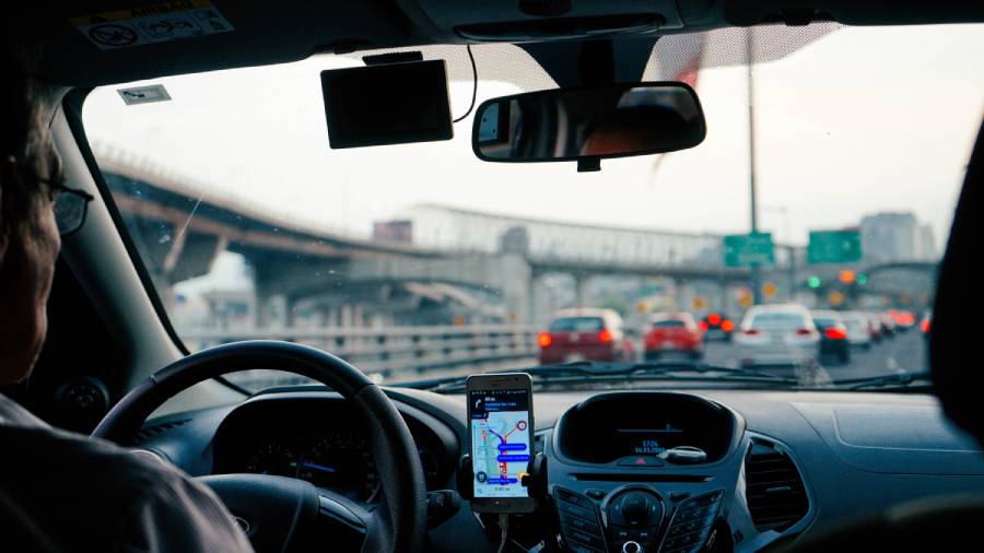 View from inside a car shows a driver's partial profile, a GPS on the dashboard, and a congested highway with blurred signs and overpasses in the background, reminiscent of navigating complex Human Resources systems in people management.