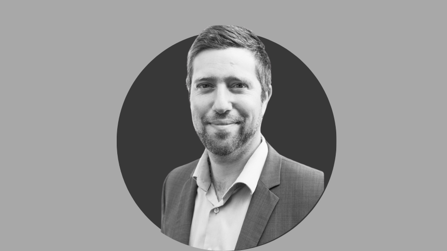 Black and white portrait of a man wearing a suit, against a circular dark background. He is smiling slightly and looking directly at the camera, perhaps reflecting on the culture while remote working.