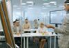 Lack of time leaves workplace learning ‘squeezed out’ Staff take part in a learning session, seated at desks while an instructor talks at a board in front.