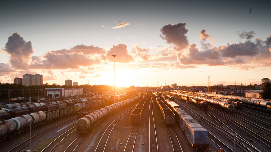 A railway yard during sunset, featuring multiple tracks with stationary trains and cargo, surrounded by buildings and a cloudy sky, where personnel diligently oversee operations to ensure everything runs smoothly.