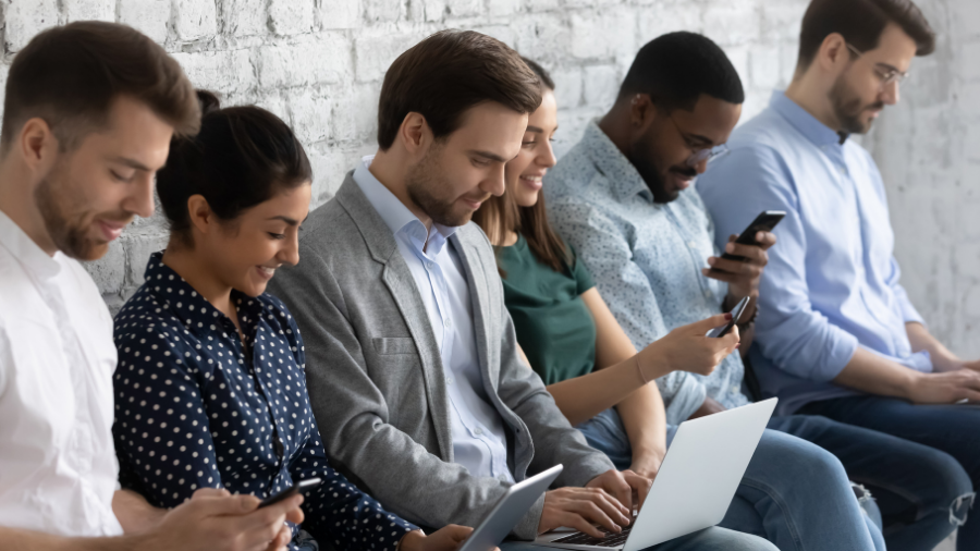A diverse group of six people sits in a row against a white brick wall, using laptops and smartphones. Two women and four men appear fully engaged with their devices, showcasing an inclusive environment.