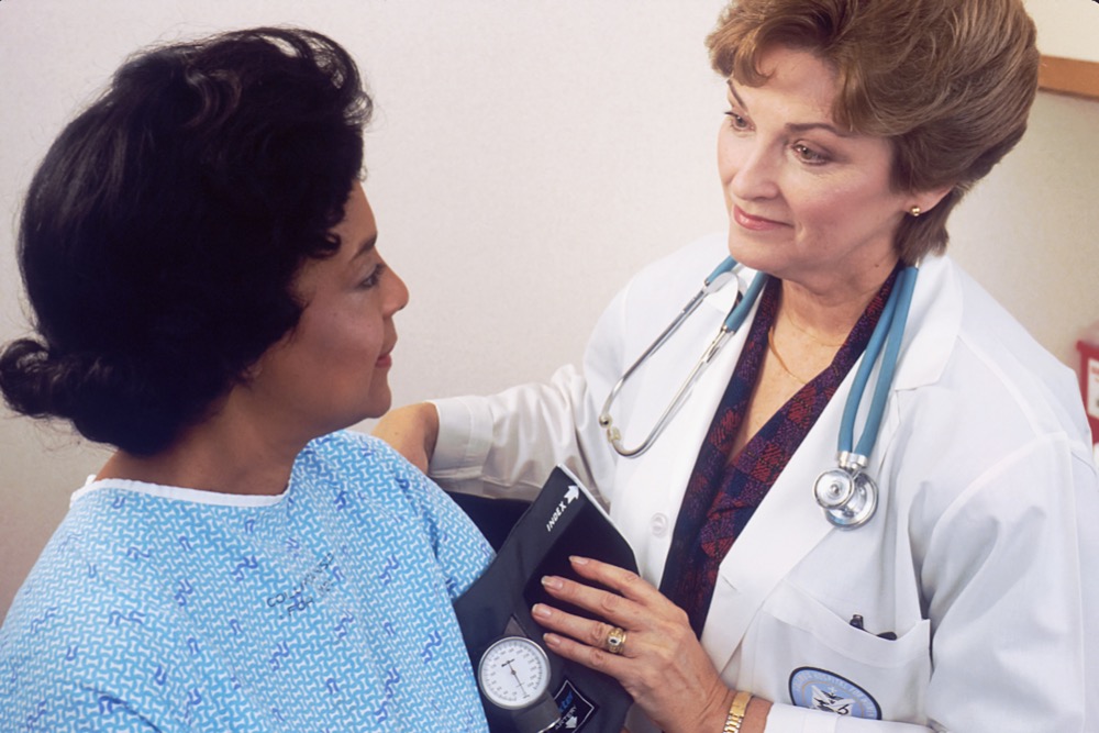 A doctor, exemplifying the vital role of women in medicine, wears a white coat and stethoscope while taking the blood pressure of a patient sitting in a hospital gown.