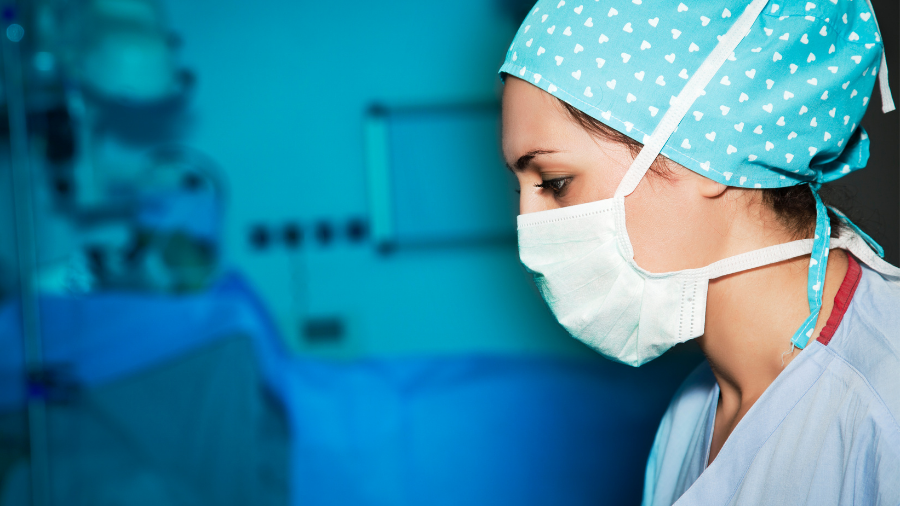 A healthcare worker wearing a surgical mask and blue scrubs looks to their right in a clinical setting within the NHS, where dedication and passion for patient care continue to increase.