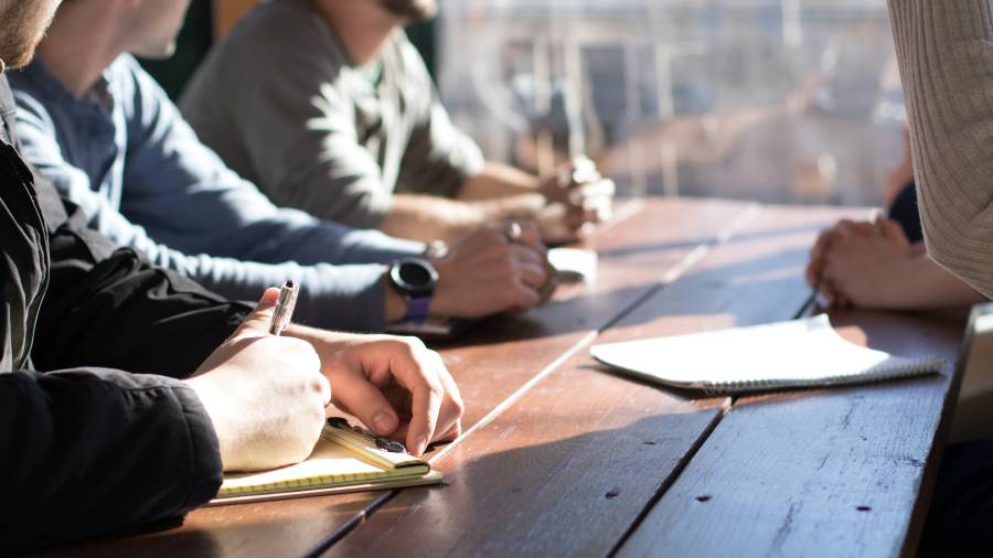 A group of individuals, including HR personnel, is sitting at a wooden table, writing in notepads and using electronic devices during a meeting.