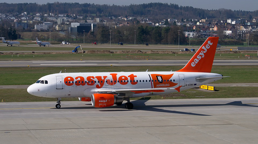 An EasyJet airplane parked on an airport runway, with buildings and trees visible in the background. The aircraft has the airline's logo and the name 