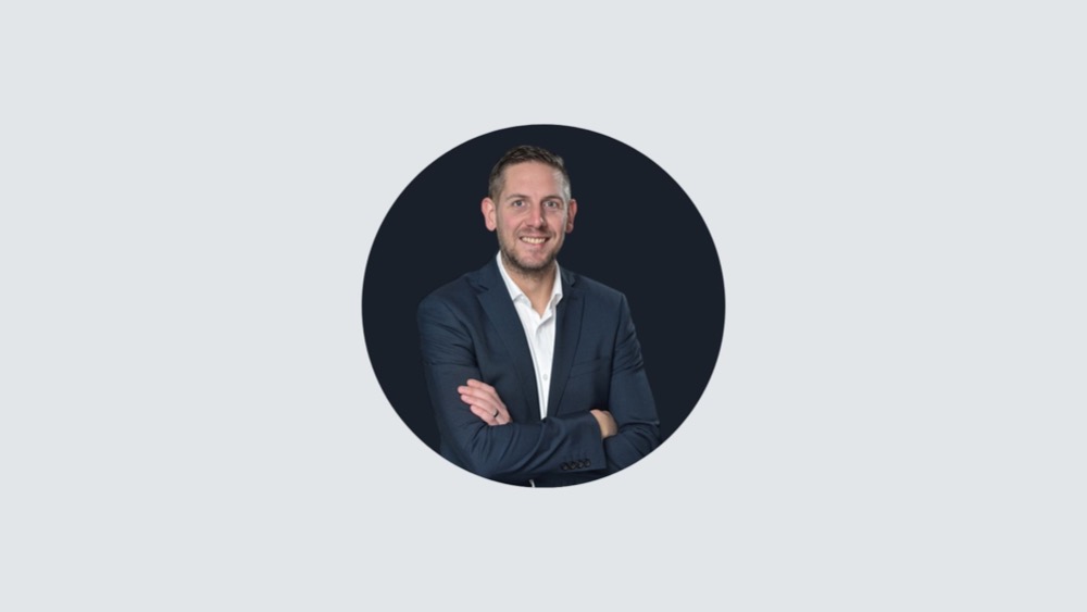 A man in a business suit poses against a dark circular background, his arms folded and smiling with confidence. His posture exudes professionalism as he navigates the complexities of IR35 regulations.