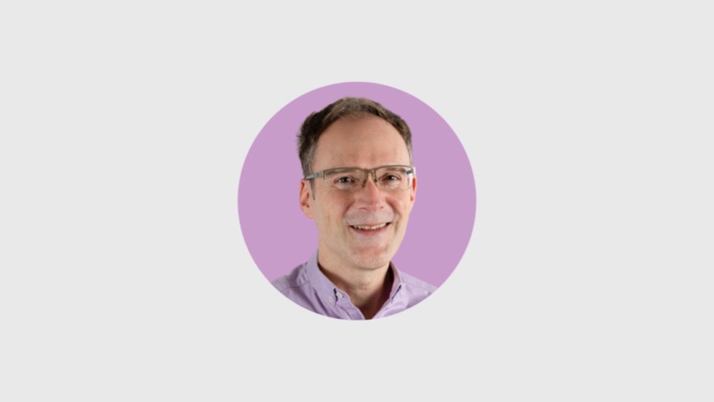A smiling man wearing glasses and a light purple shirt is positioned in front of a purple circular background, embodying optimism amidst the recruitment crisis.