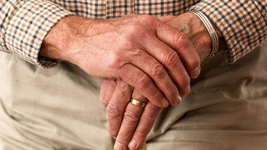 Close-up of elderly hands resting on a cane, one hand wearing a gold ring. The individual, with years of personnel experience, is dressed in a checkered shirt and beige pants.
