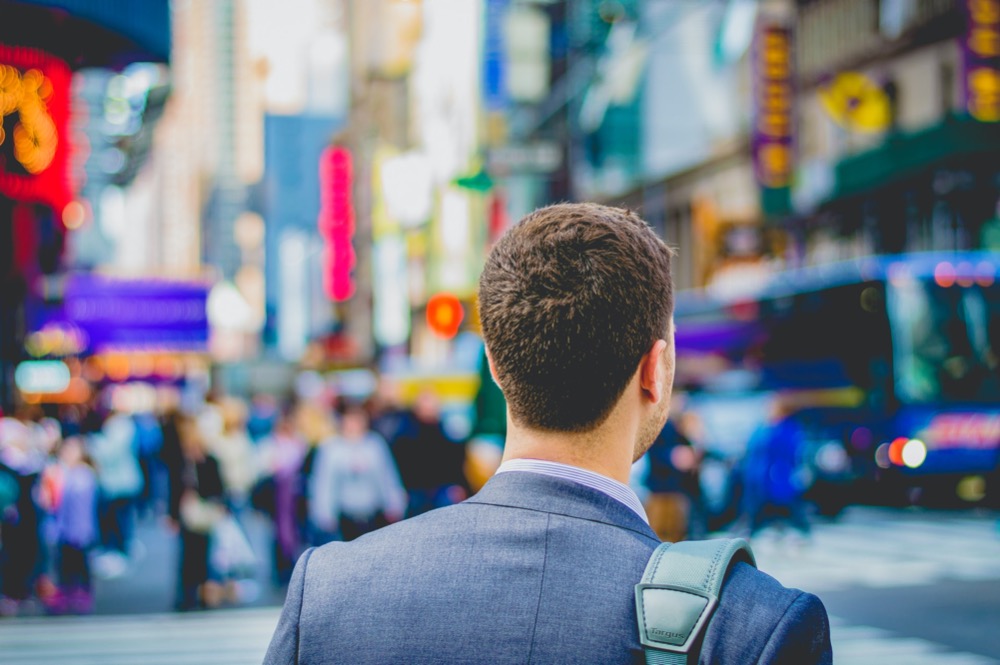 A man in a suit stands with his back to the camera in a busy cityscape, colorful lights and blurred pedestrians all around him, reflecting the hustle and bustle reminiscent of current recruitment trends.
