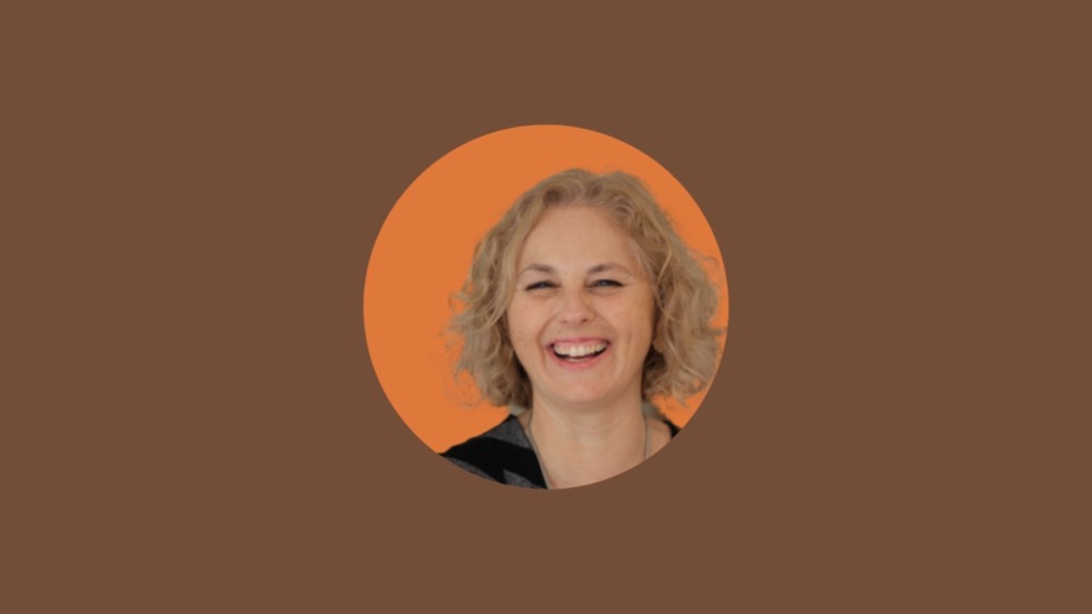A woman with short blonde hair is smiling, set against an orange circle on a brown backdrop. Her confident expression subtly hints at her advocacy for closing the gender pay gap.