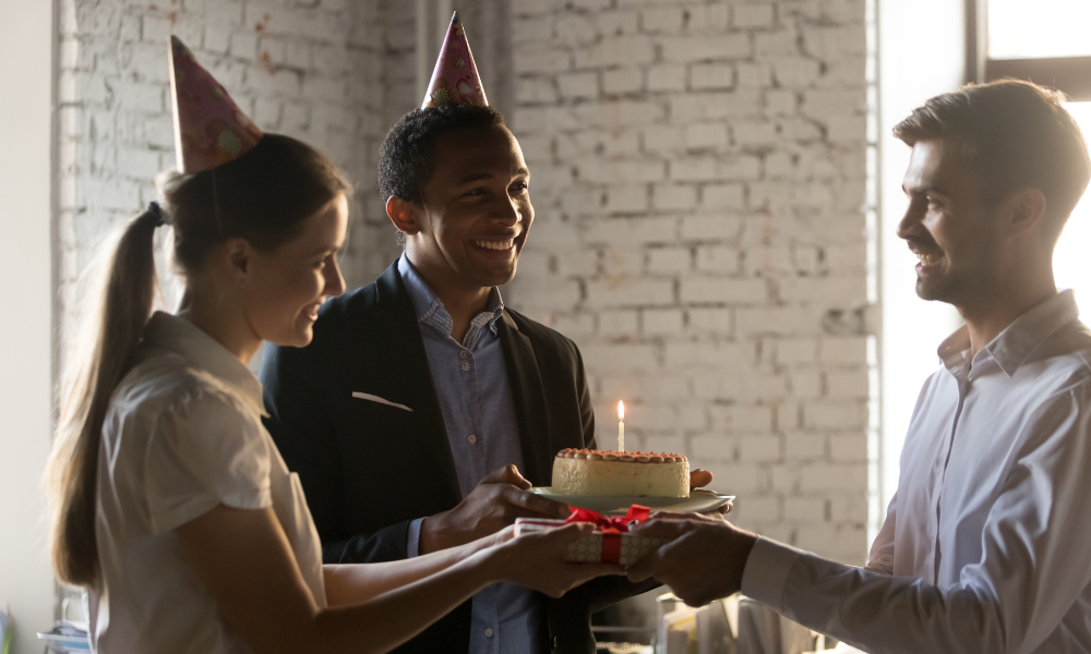 Three people wearing party hats, with one holding a birthday cake with a lit candle and the other two holding a gift. They appear to be celebrating Employee Appreciation Day in a room with a brick wall background.