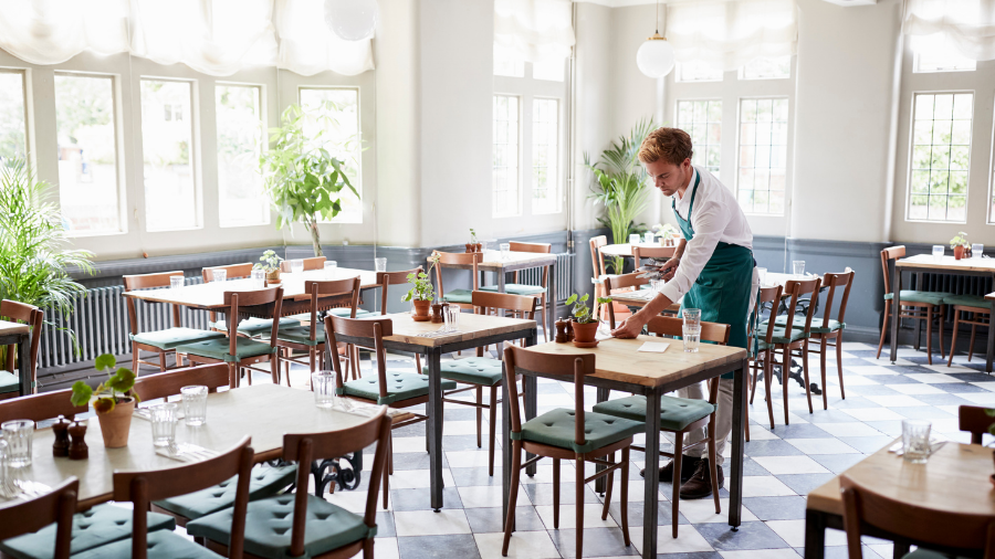 A waiter arranges items on a table in a bright, empty restaurant with wooden chairs, checkered floor tiles, and potted plants by the windows, reflecting the omicron uncertainty that has kept patrons away.