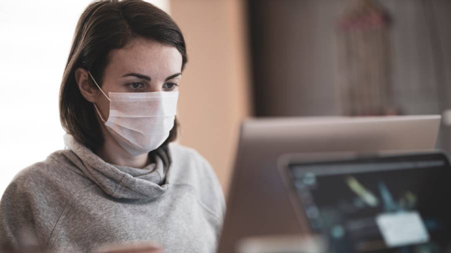 A person wearing a face mask and a grey hoodie is sitting indoors, looking at a laptop screen, perhaps reviewing important HR documents.