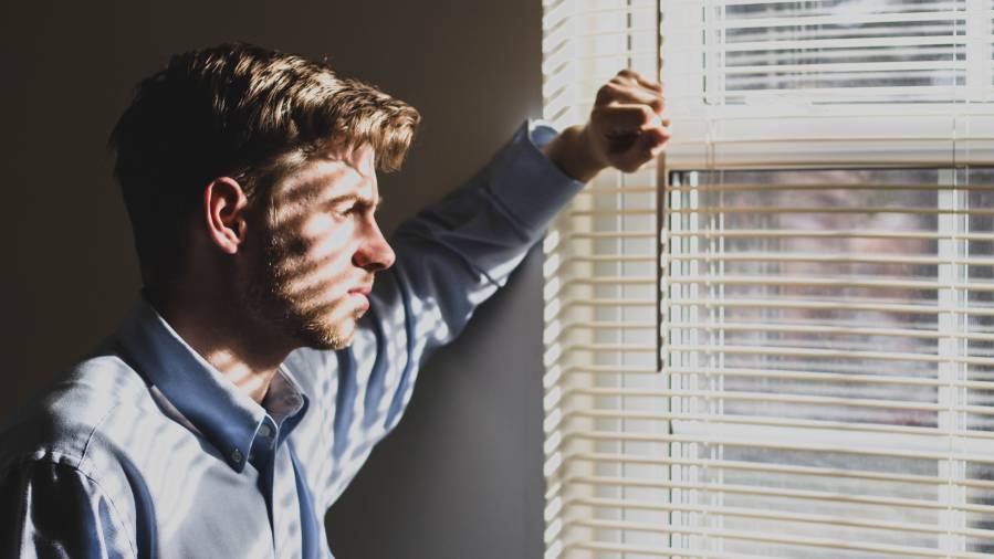 A man in a blue shirt stands near a window with blinds, his face partially illuminated by sunlight filtering through the slats, looking contemplative, possibly pondering an HR decision.