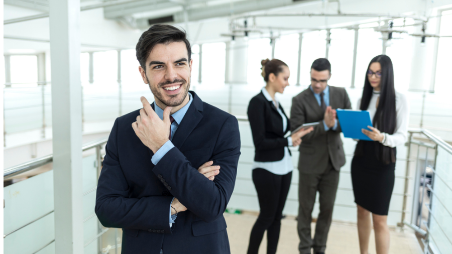 A man in a suit and tie stands smiling in the foreground. In the background, three people are engaged in a discussion, holding documents and a tablet, emphasizing collaboration without ethnicity bias.