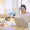 Three women sitting at a table in an office meeting. One woman on the left is shaking hands with a woman in the center, showcasing effective People Management. A laptop, documents, and glasses of water are on the table.