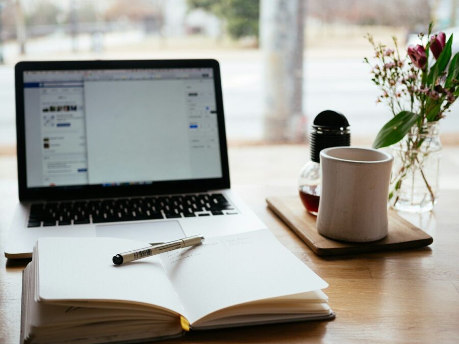 A laptop and notebook and pen beside a mug and flowers in a home office setup.