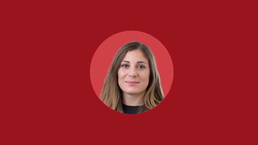 Portrait of a woman with long brown hair and a slight smile set against a solid red background, capturing the essence of confidence often sought in job candidates.