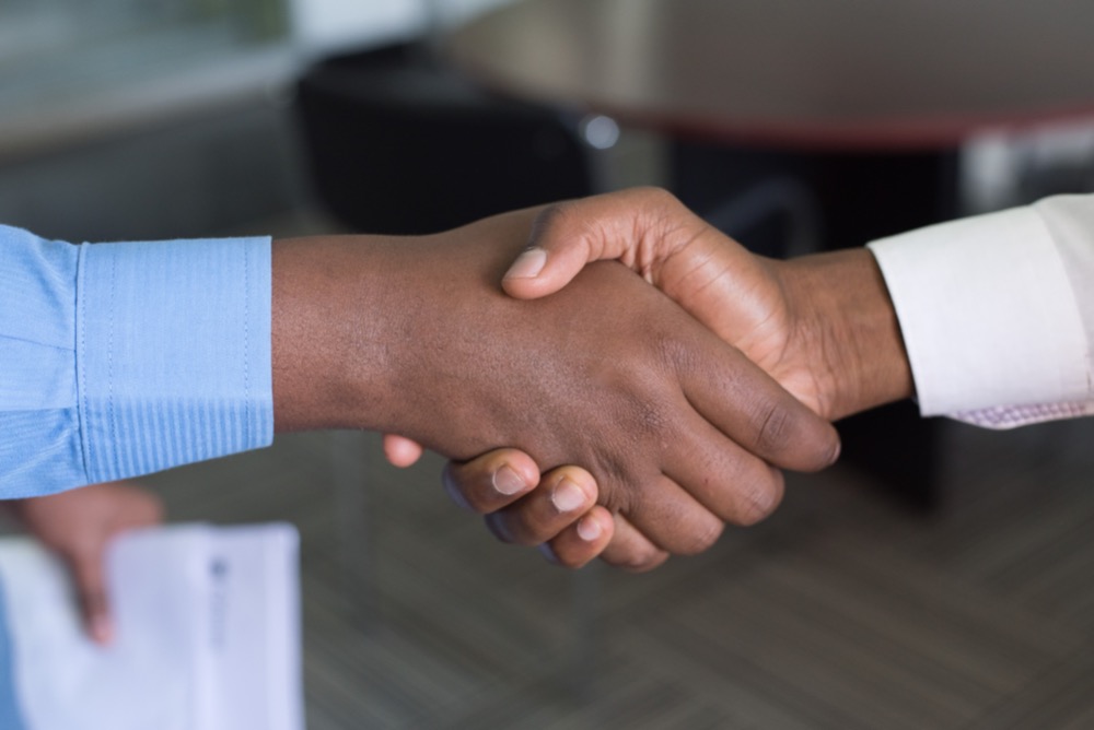 Two people are shaking hands, with one person holding a piece of paper—a classic scene of M&A deals. The background shows an office setting with a chair and a table.