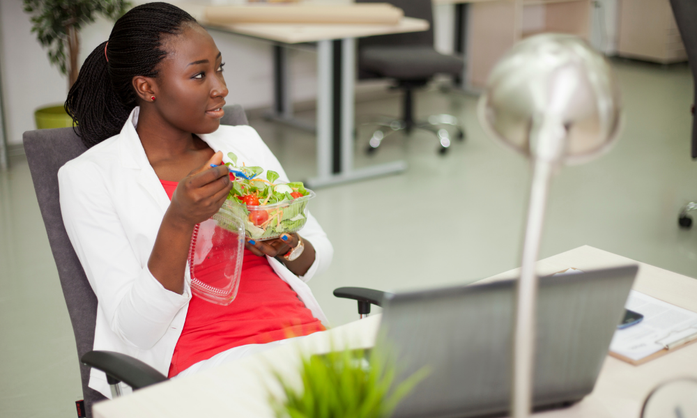 A black woman in a white jacket and red top looking off camera and smiling, while eating a salad at her desk