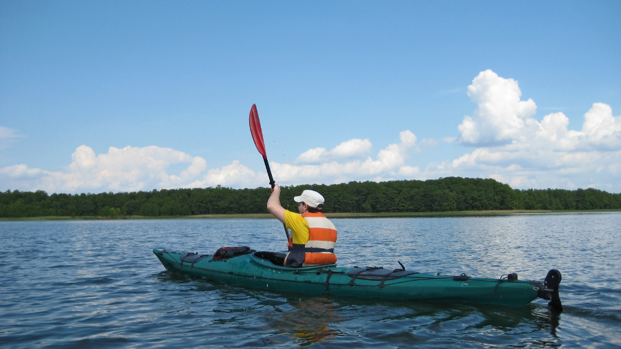 A person wearing a life jacket paddles a green kayak on a calm lake with a forested shoreline, enjoying the serenity and scattered clouds, perhaps contemplating the benefits of a four day week.