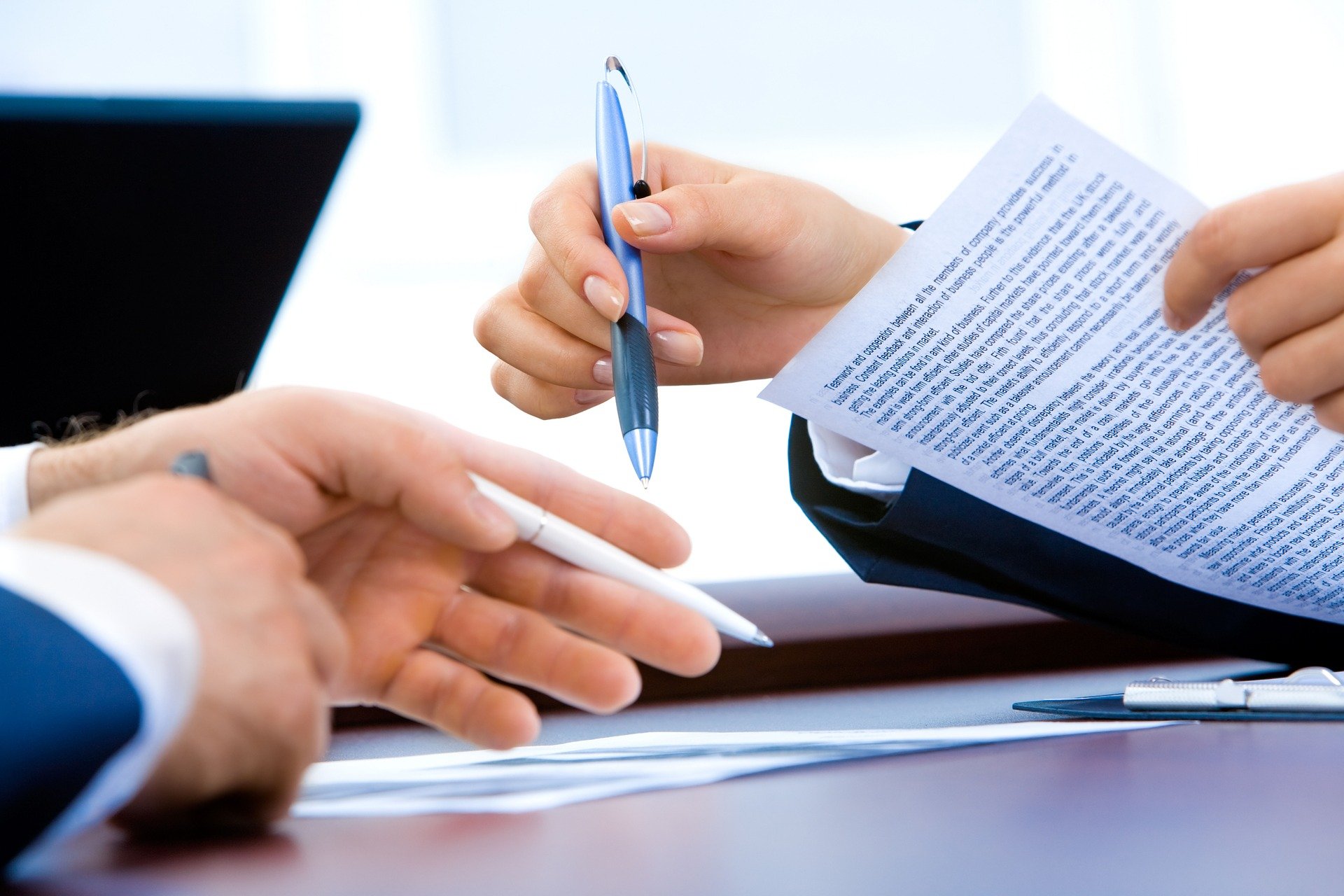Two people in business attire holding pens, one also holding a document with printed text under IR35 regulations. The scene suggests a discussion or signing of the document. A laptop is visible in the background.