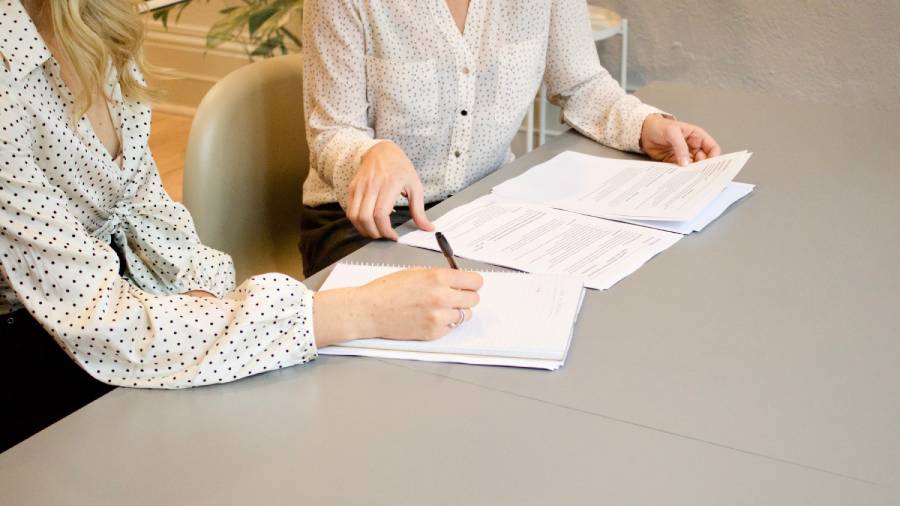 Two people sit at a gray table, one writing in a notebook and the other pointing at documents. Both are wearing white shirts with black dot patterns, discussing HR strategies. Papers and a pen are on the table, emphasizing their focus on effective People Management.