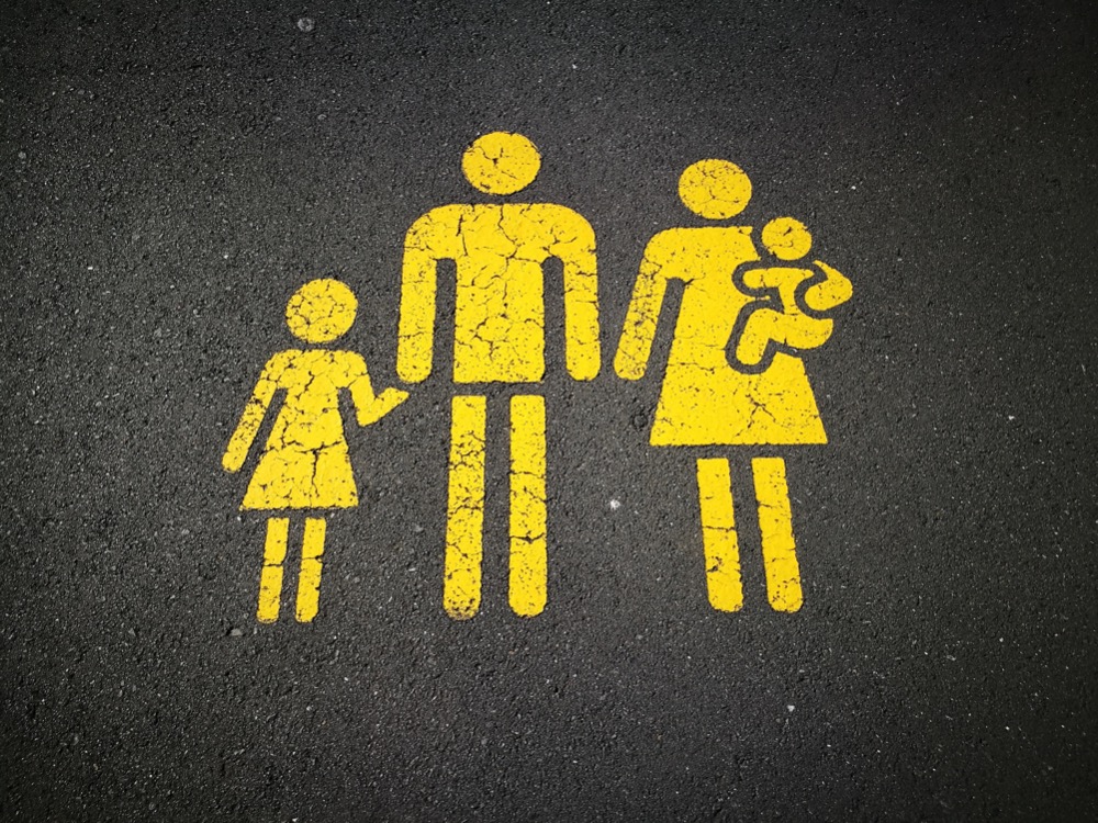 Yellow painted symbols of a family, with a child, an adult holding a baby, and another adult on a dark asphalt surface subtly highlight societal issues like the gender pay gap.