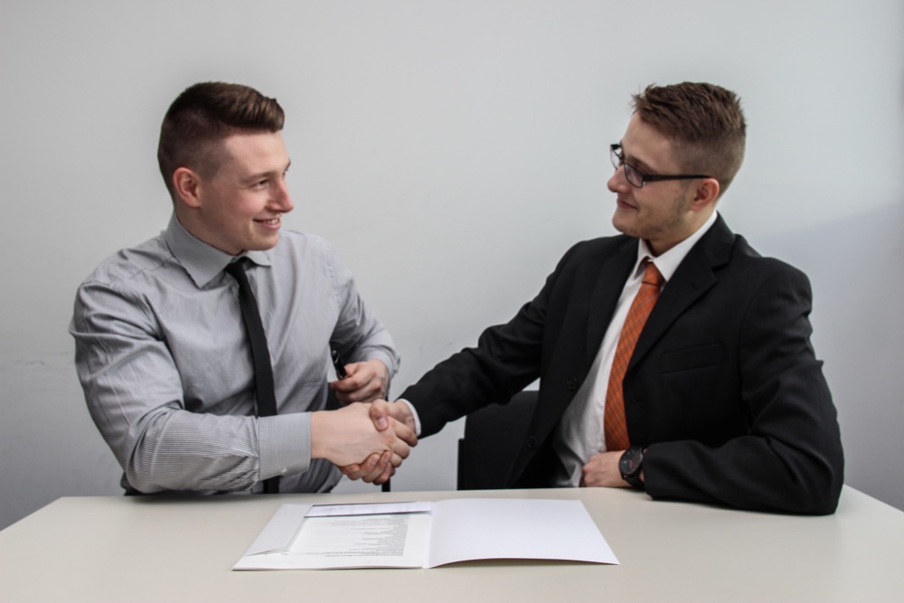Two men in business attire shake hands across a table with documents placed on it, suggesting a successful agreement or meeting to address the skills gap.