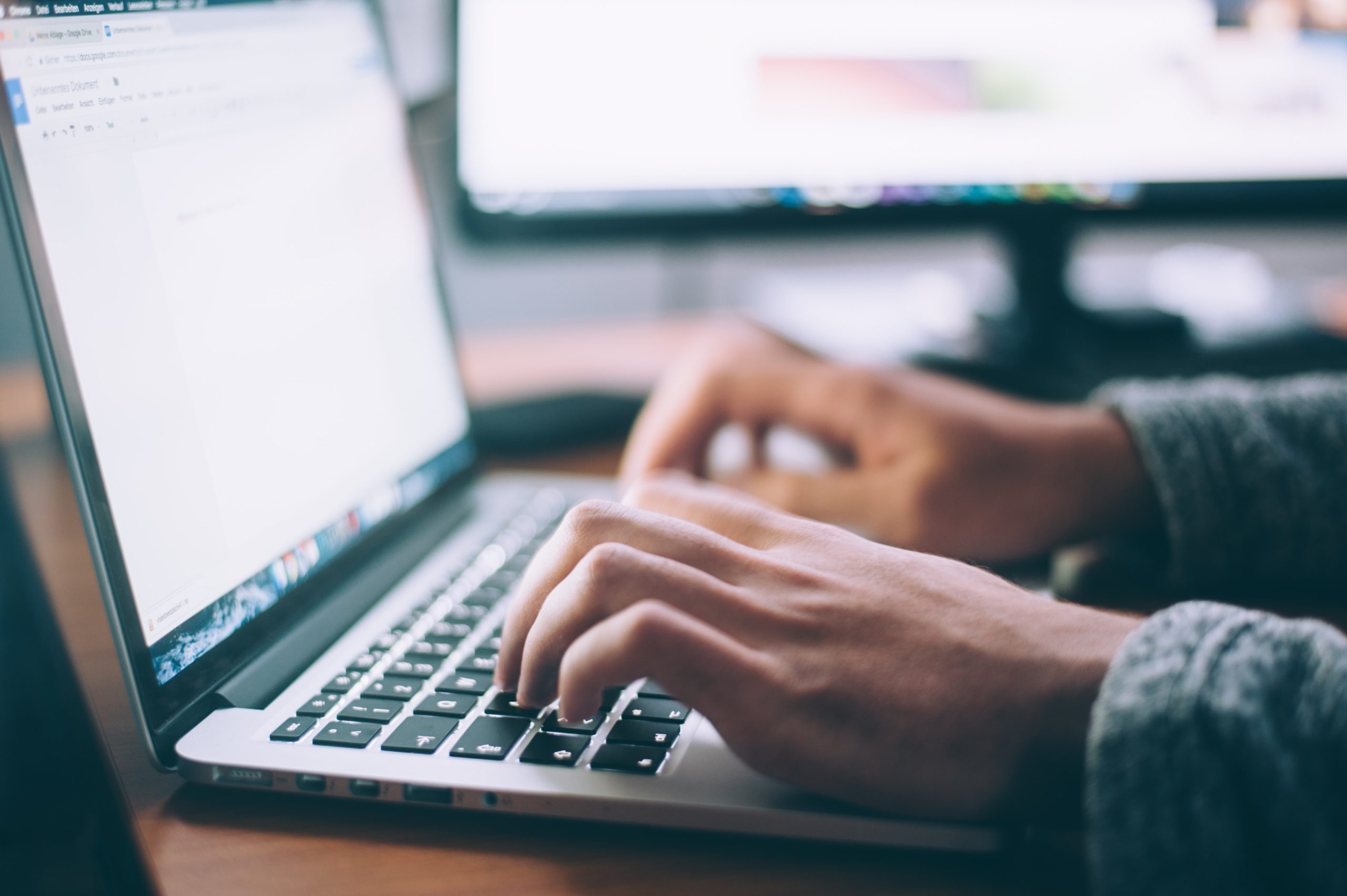 Person typing on a laptop keyboard with two computer monitors in the background, possibly working on critical whistleblowing documents.