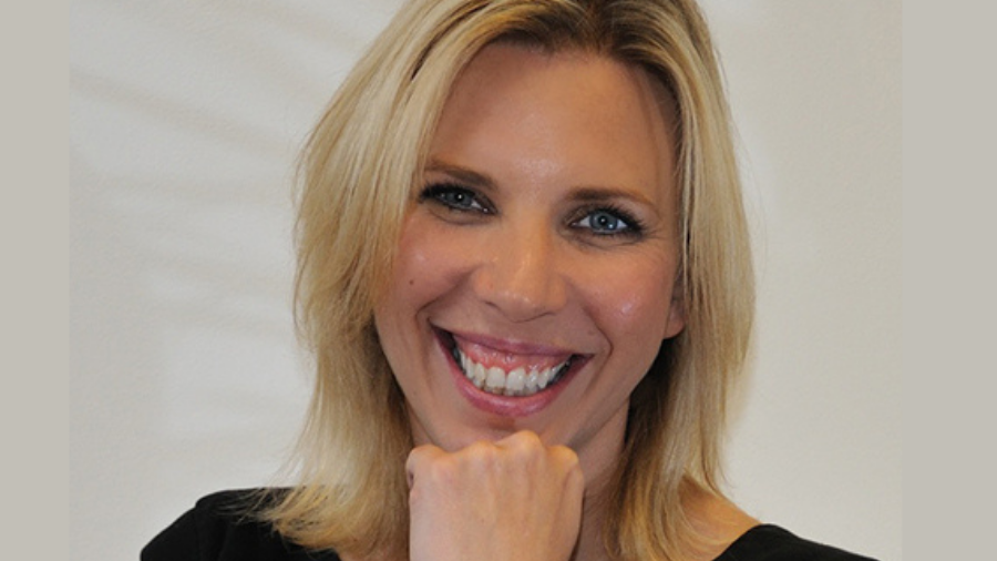 A smiling person with shoulder-length blonde hair and light skin, resting their chin on their hand, epitomizes the essence of wellbeing amid remote working, against a plain background.